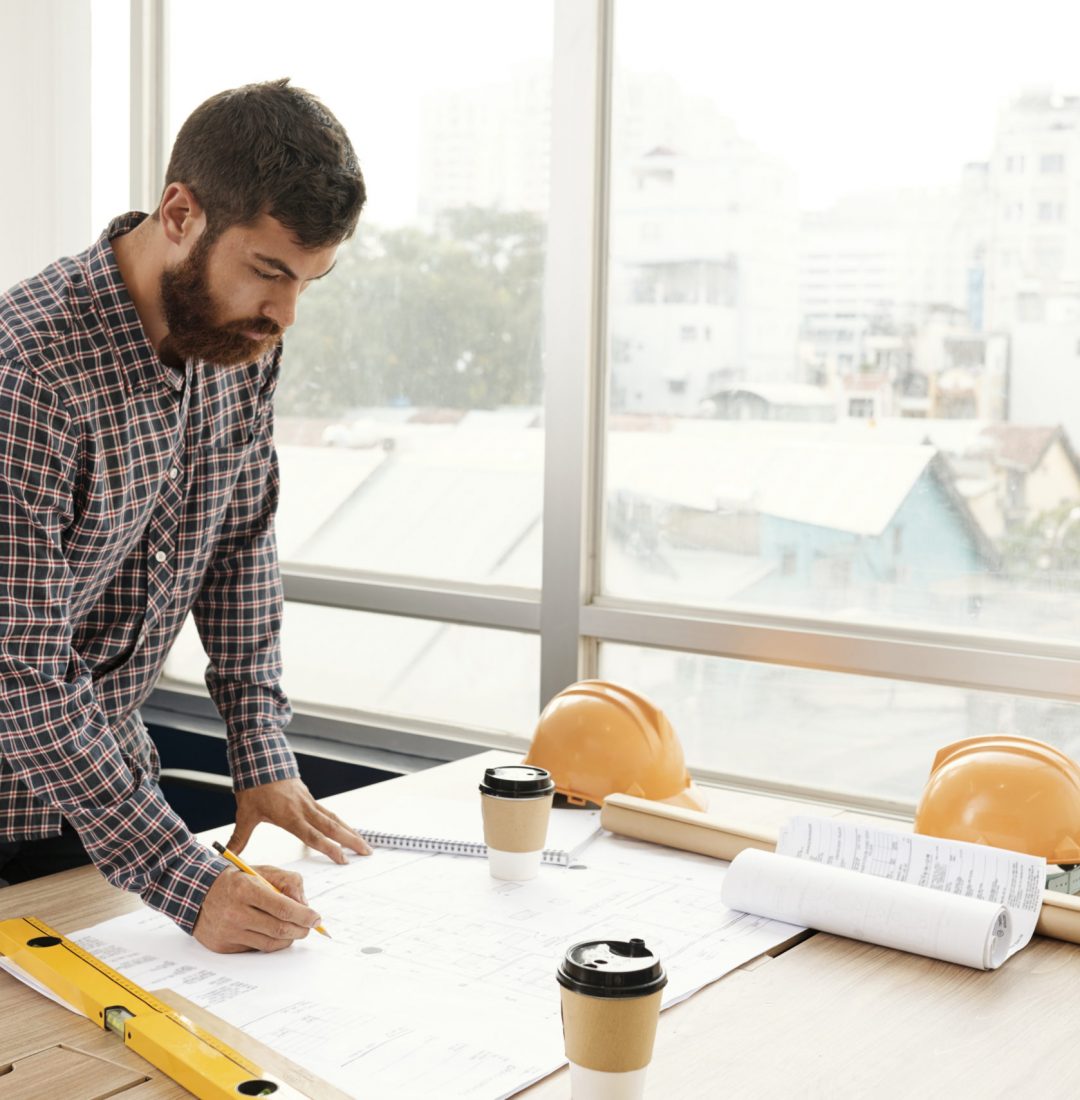 Young bearded engineer in casual clothing standing near his workplace and drawing construction blueprint with pencil at office